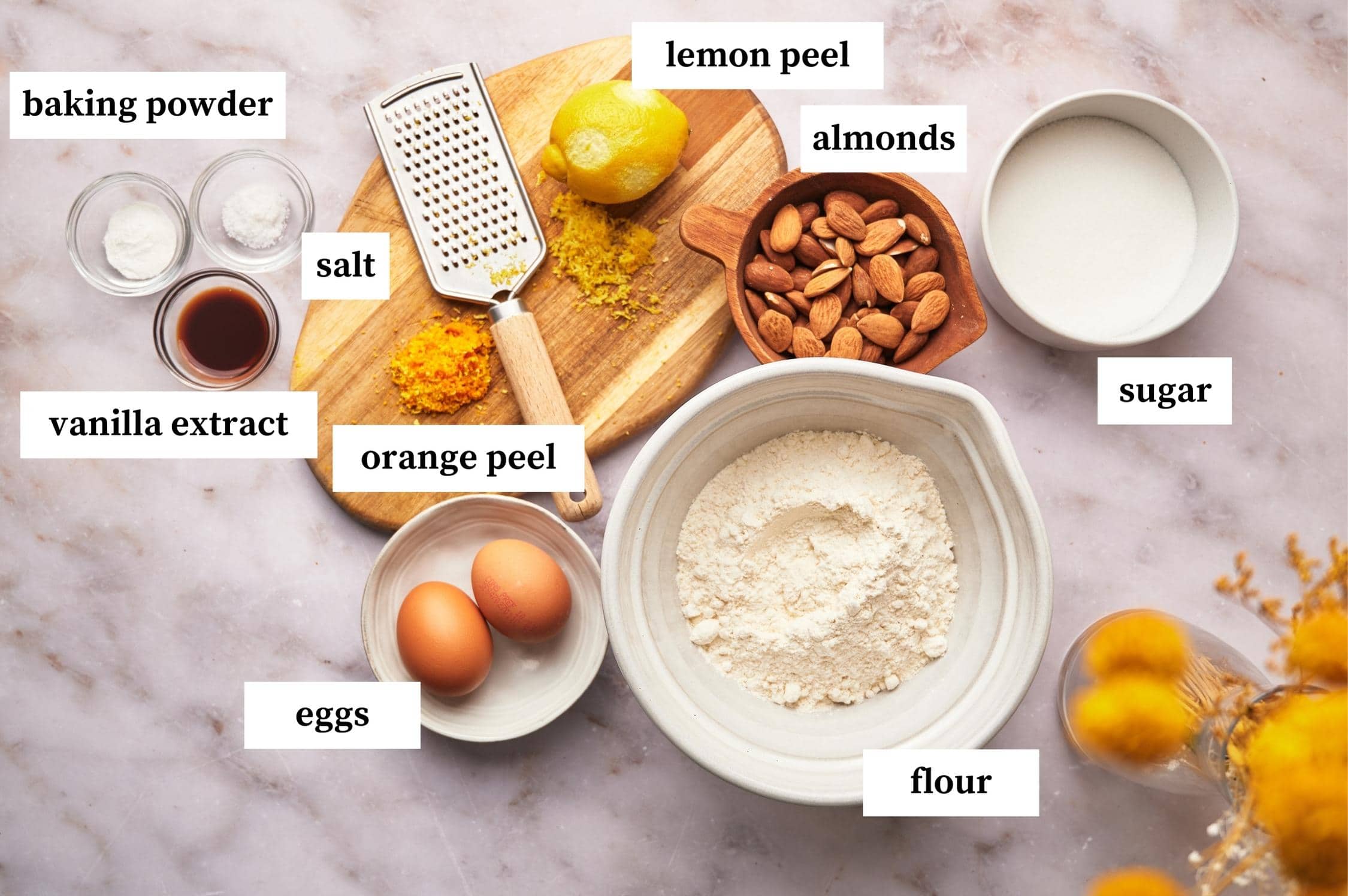Ingredients for biscotti recipe on a table: flour, eggs, sugar, almonds, lemon peel, orange peel, baking powder, salt and vanilla extract
