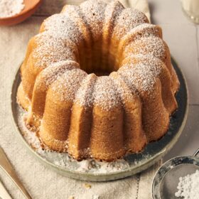 ciambella italian cake dusted with powdered sugar on a plate sitting on top of a table mat. a glass of milk, forks, a small bowl and a small sifter with powdered sugar on it surround the plate.