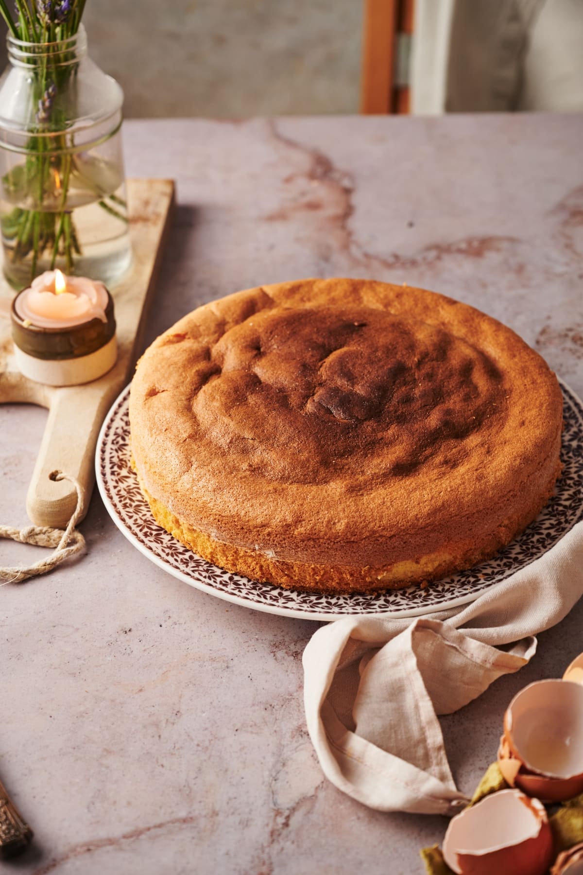baked italian sponge cake on a floral plate on top of a marble counter top next to a wooden chopping board with a candle and vase of flowers.