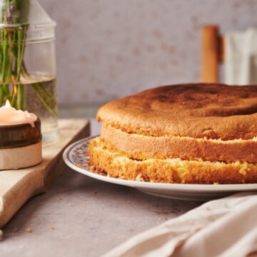 italian sponge cake cut into three layers on a plate on top of a marble counter top with a candle and a vase of flowers on top of a wooden chopping board.