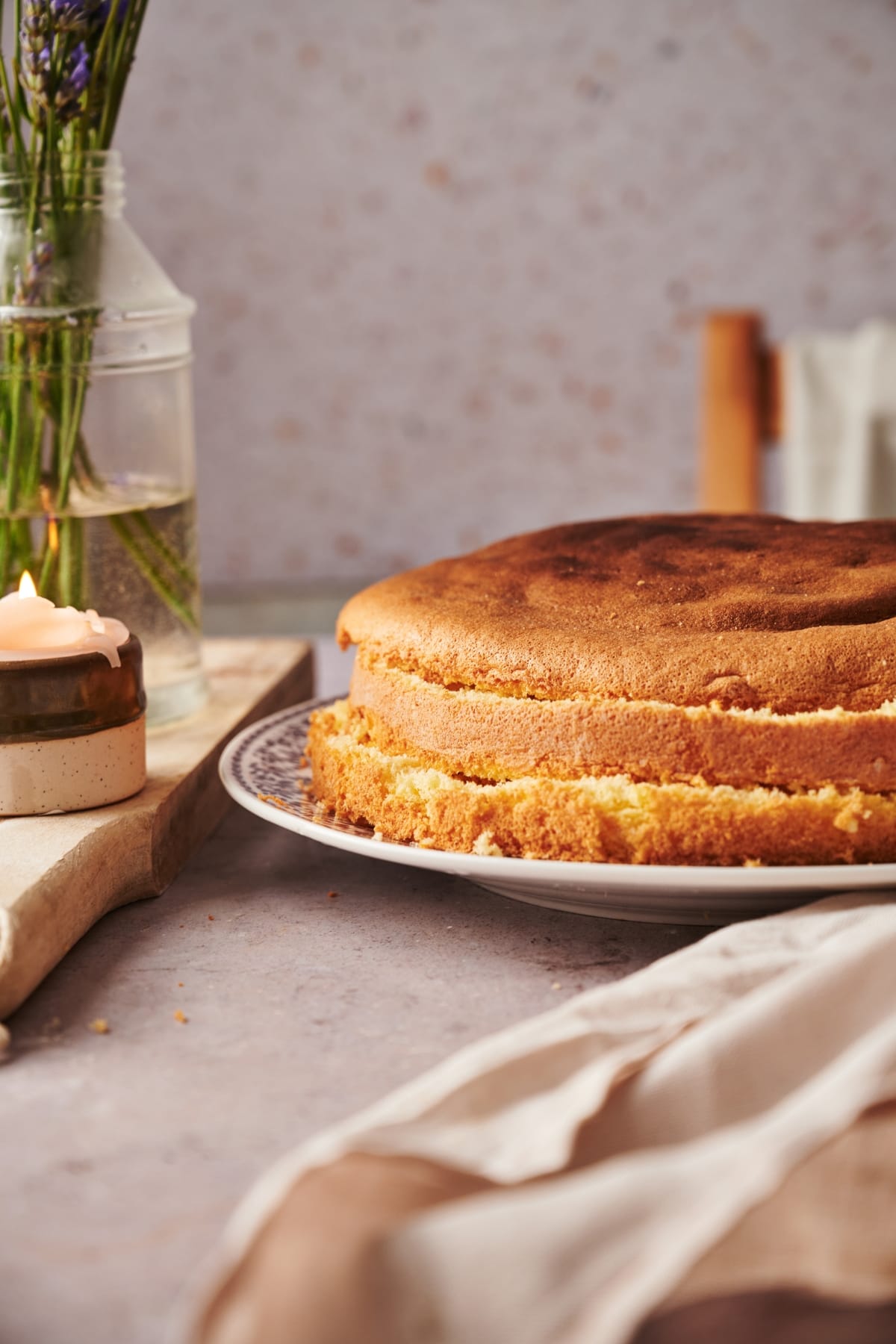 italian sponge cake cut into three layers on a plate on top of a marble counter top with a candle and a vase of flowers on top of a wooden chopping board.