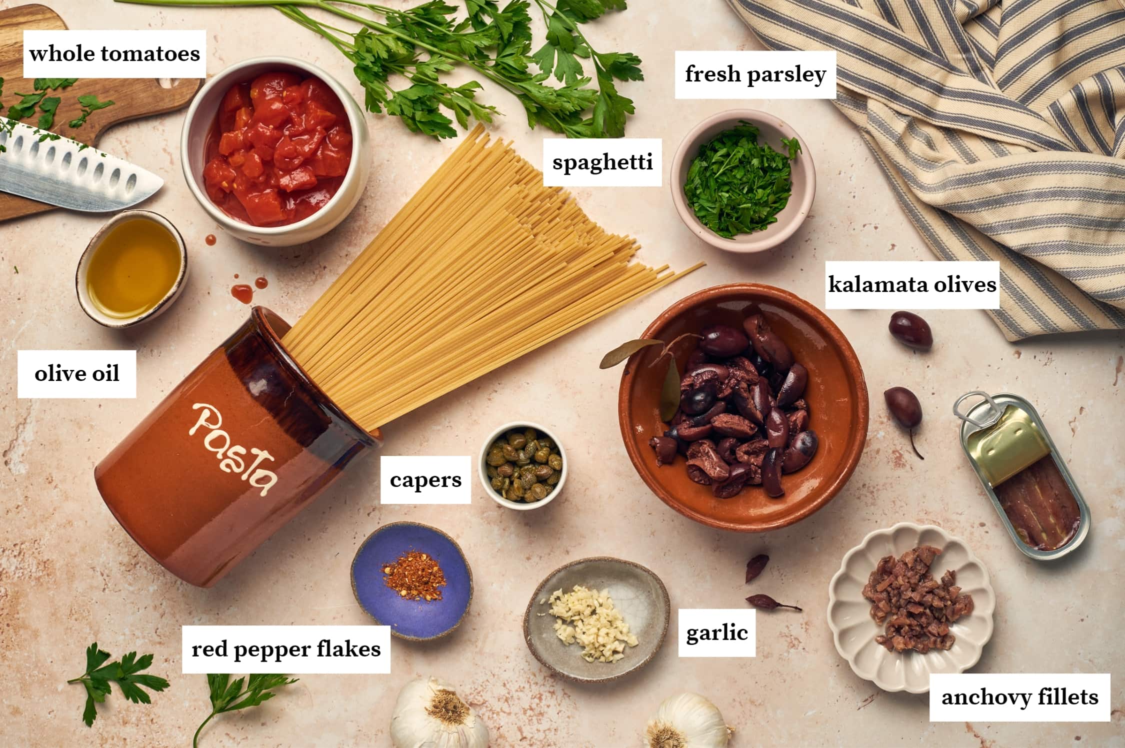 Ingredients for pasta puttanesca on a table: spaghetti pasta, tomatoes, fresh parsley, kalamata olives, garlic, anchovy fillets, garlic, red pepper flakes, olive oil.