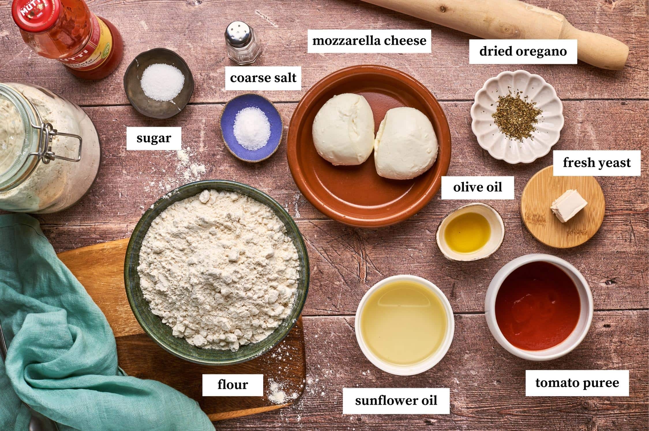 ingredients for panzerotti recipe on a table: flour, sunflower oil, tomato puree, fresh yeast, dried oregano, olive oil, mozzarella cheese, coarse salt, sugar.