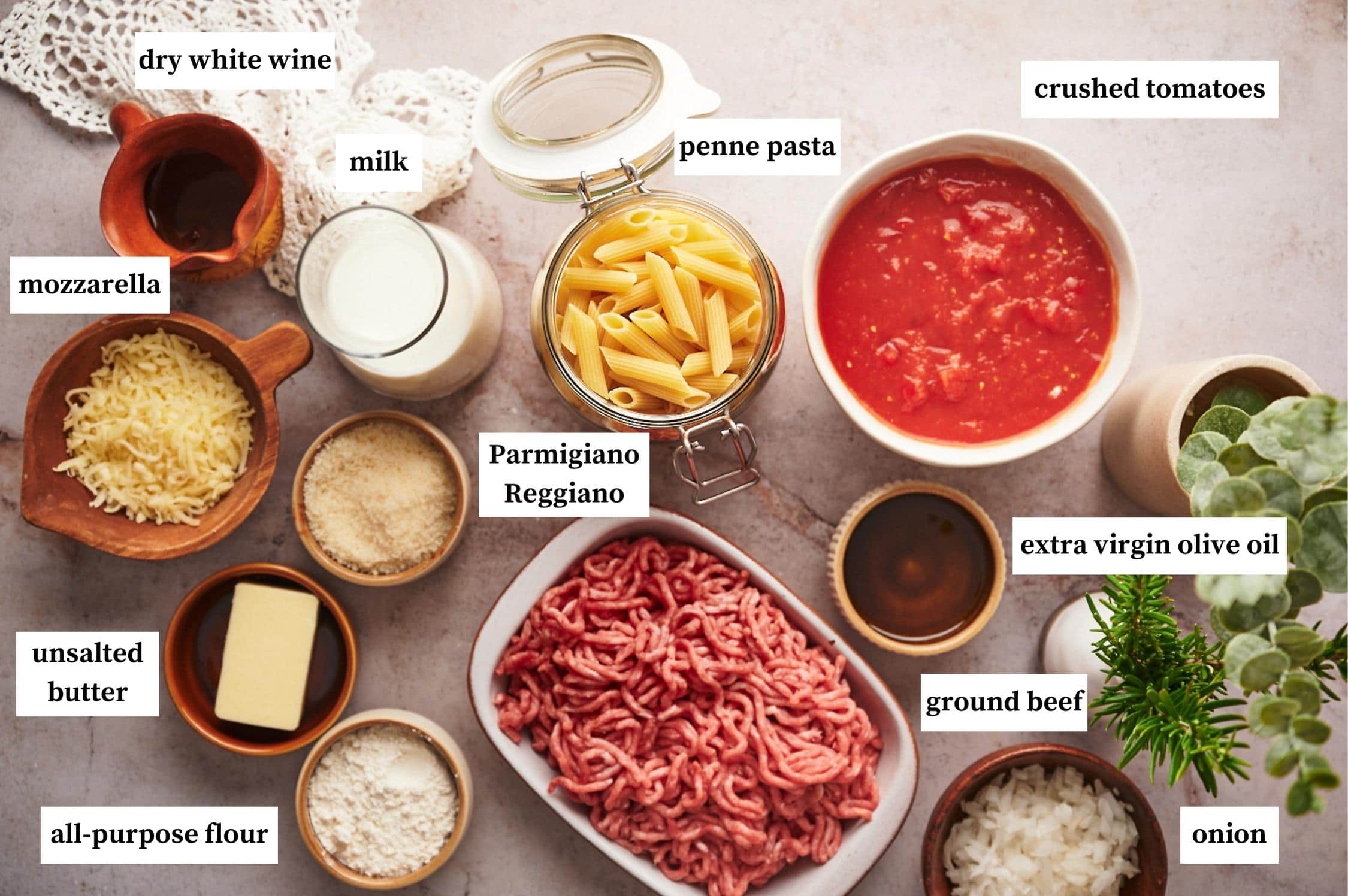 baked penne ingredients: small pitcher of dry white wine, jar of milk, jar of penne pasta, bowl of crushed tomatoes, bowl of mozzarella, parmigiano reggiano, unsalted butter, extra virgin olive oil, rectangular dish with ground beef in it, bowl of all-purpose flour and onions.