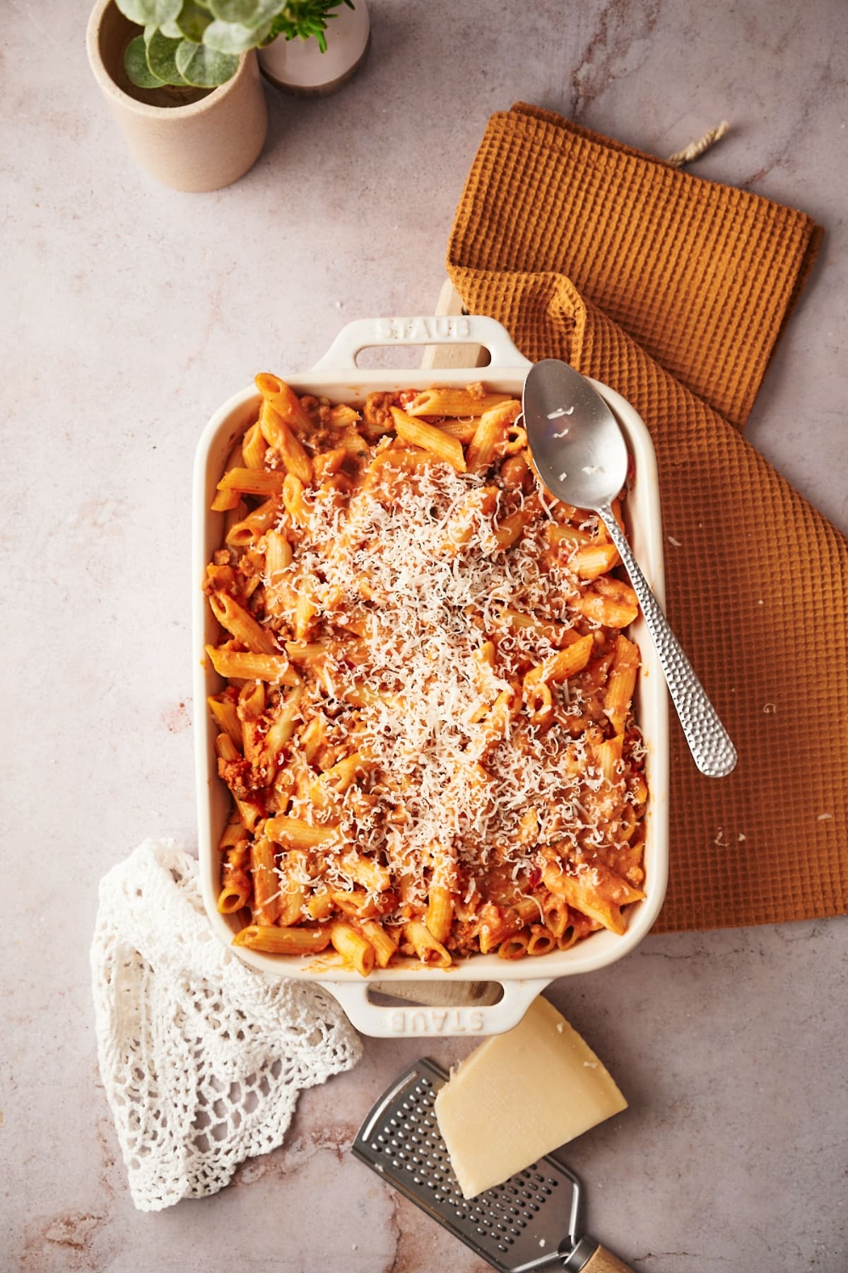 baked penne in a white casserole dish with a spoon on it. an orange table napkin, white lace and cheese and grater around the dish.
