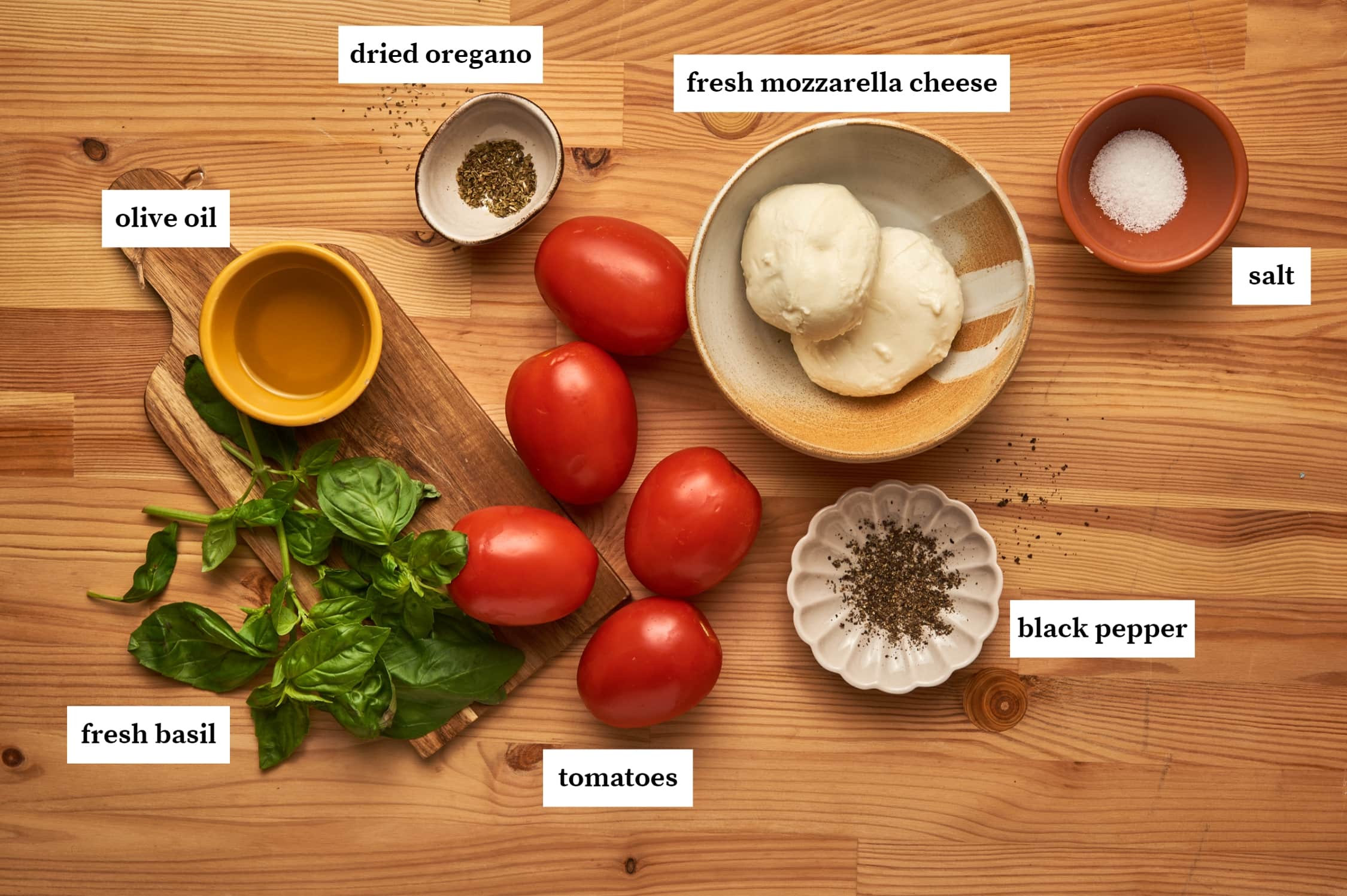 ingredients of caprese salad: fresh basil and a bowl of olive oil on top of a wooden chopping board. some tomatoes on top of a wooden counter top next to a bowl of dried oregano, fresh mozzarella cheese, salt and ground black pepper.