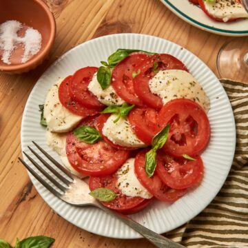 a dish of caprese salad on a white plate with a fork on top of a kitchen towel. next to a larger plate of caprese salad, bowl of salt, some wine glass.
