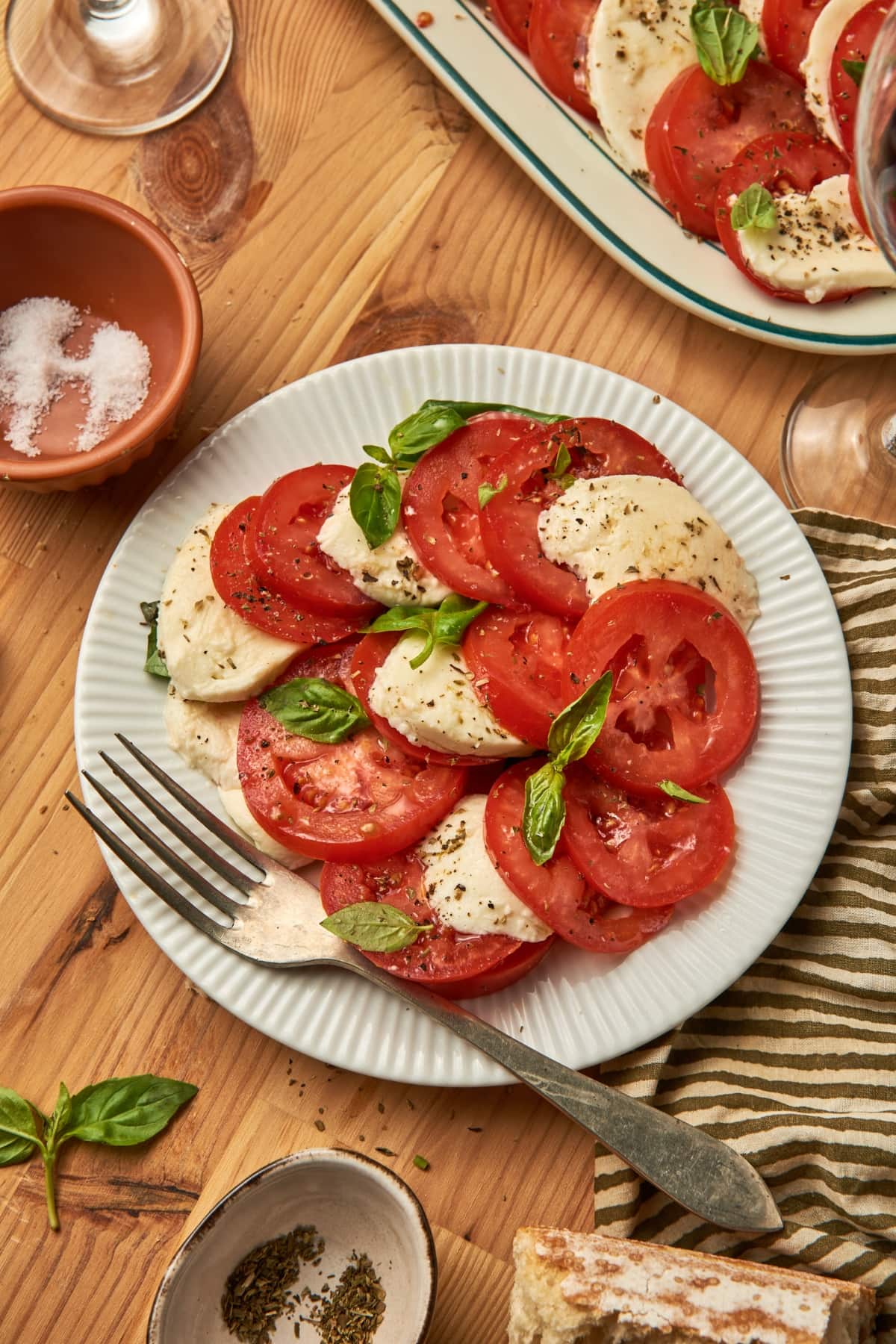 a dish of caprese salad on a white plate with a fork on top of a kitchen towel. next to a larger plate of caprese salad, bowl of salt, some wine glass.