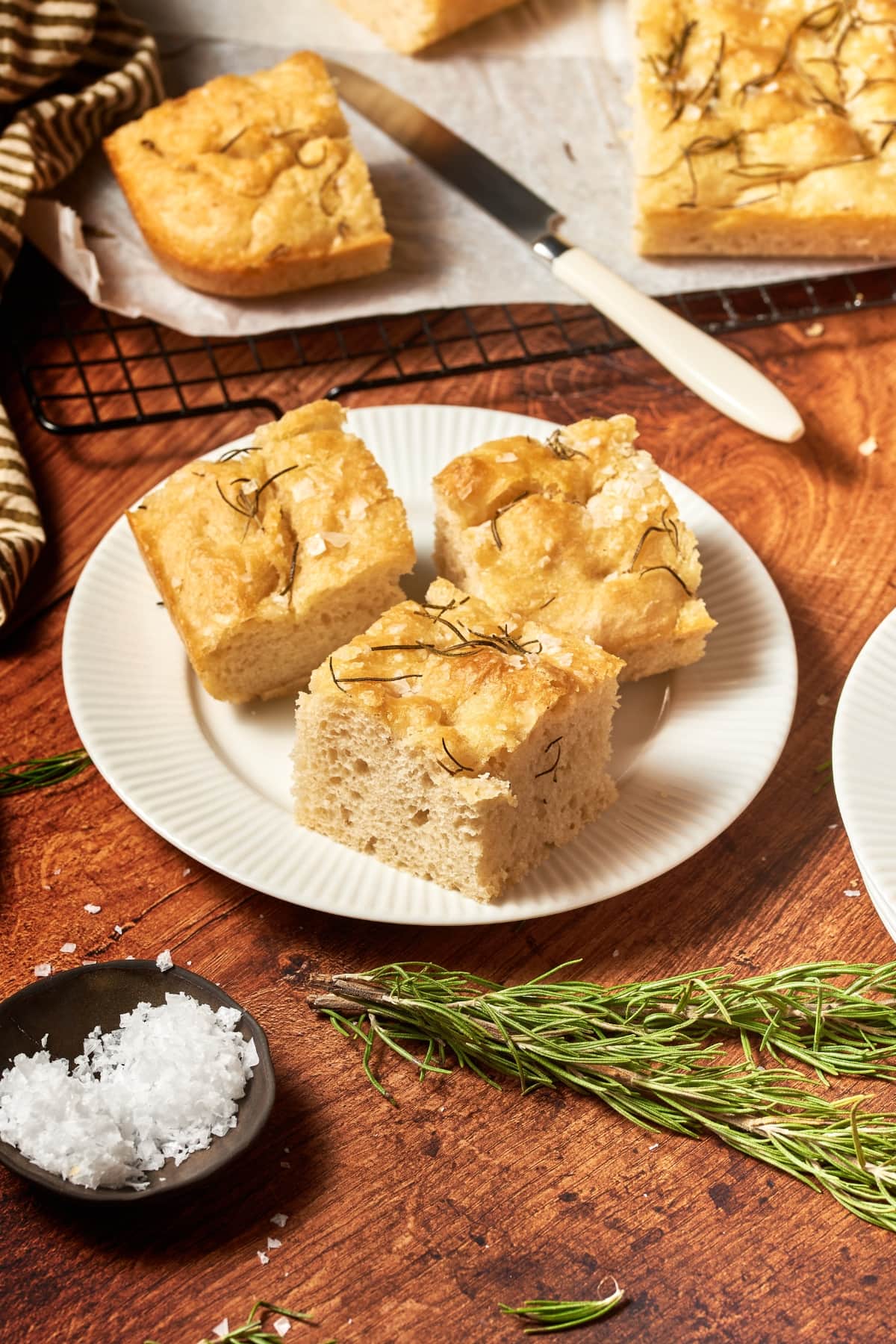 focaccia bread on a white plate with a cooling rack with more bread, some sprigs of rosemary and salt around the plate.