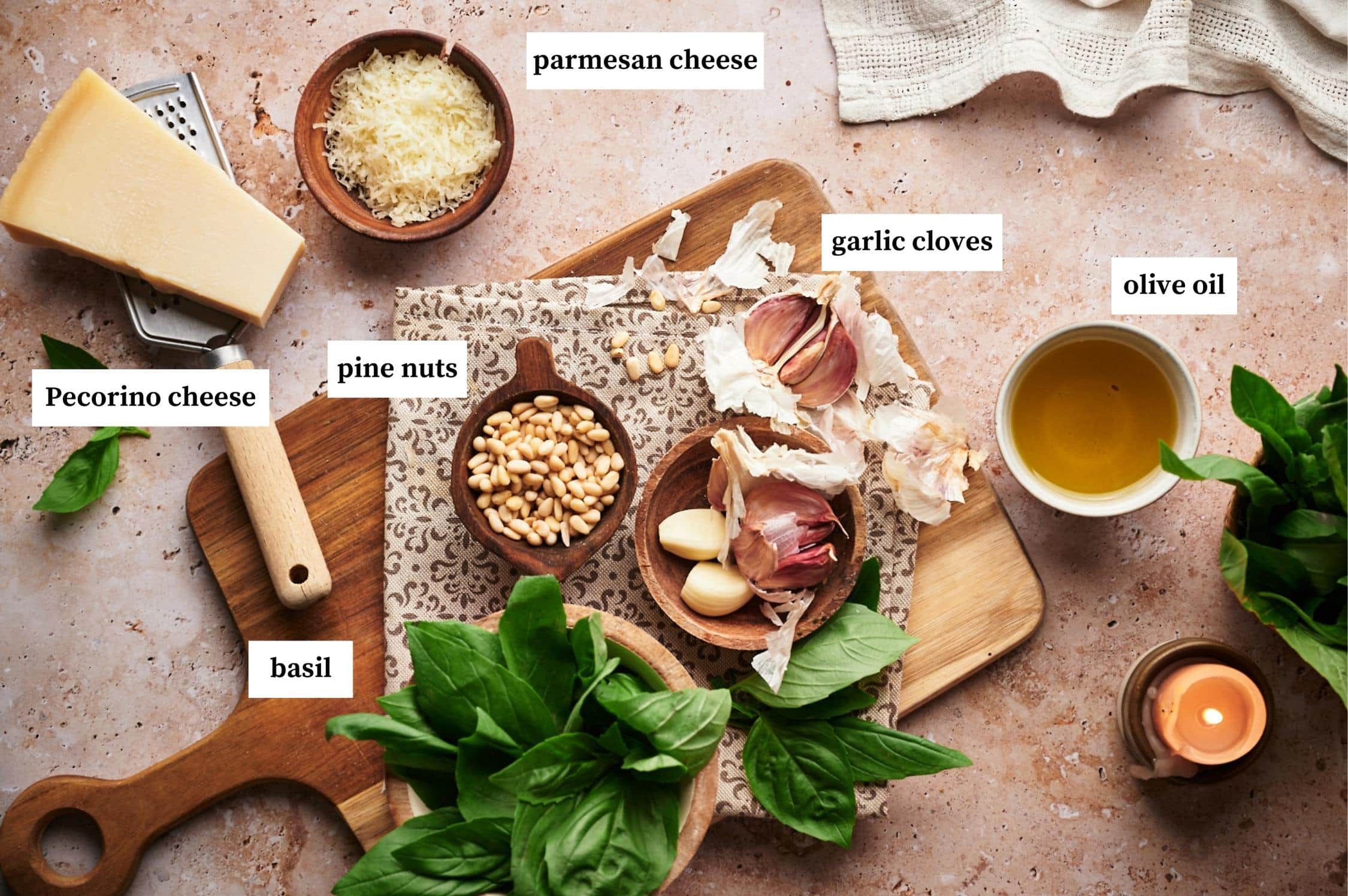 ingredients for basil pesto: bowl of parmesan cheese, pecorino cheese on top of a grater, bowl of olive oil. fresh basil leaves in a bowl, garlic cloves, pine nuts on top of a wooden chopping board.