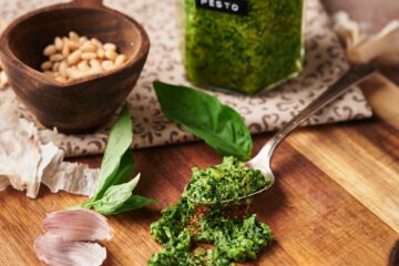 clear jar of basil pesto with a black label with white text labeled pesto. beside a pestle with pine nuts inside. some fresh basil leaves and garlic peel scattered around. and a spoon of pesto.
