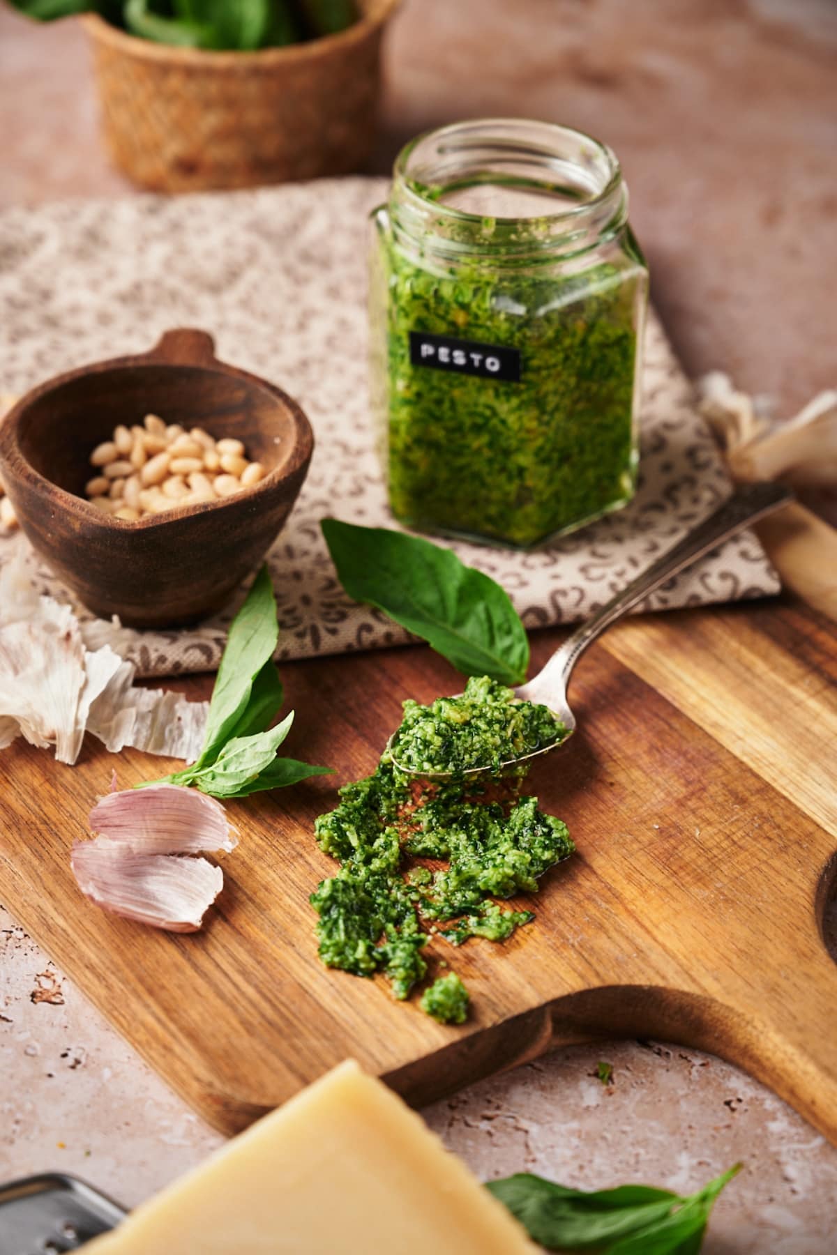 clear jar of basil pesto with a black label with white text labeled pesto. beside a pestle with pine nuts inside. some fresh basil leaves and garlic peel scattered around. and a spoon of pesto.