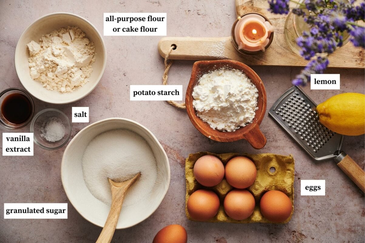 italian sponge cake ingredients, bowl of potato starch, salt and vanilla extract in separate small glass bowls, all-purpose flour in a wooden bowl with spout, large bowl of granulated sugar with a wooden spoon, six brown eggs in an egg carton, grater and a lemon, wooden chopping board with a candle and vase of lavender.