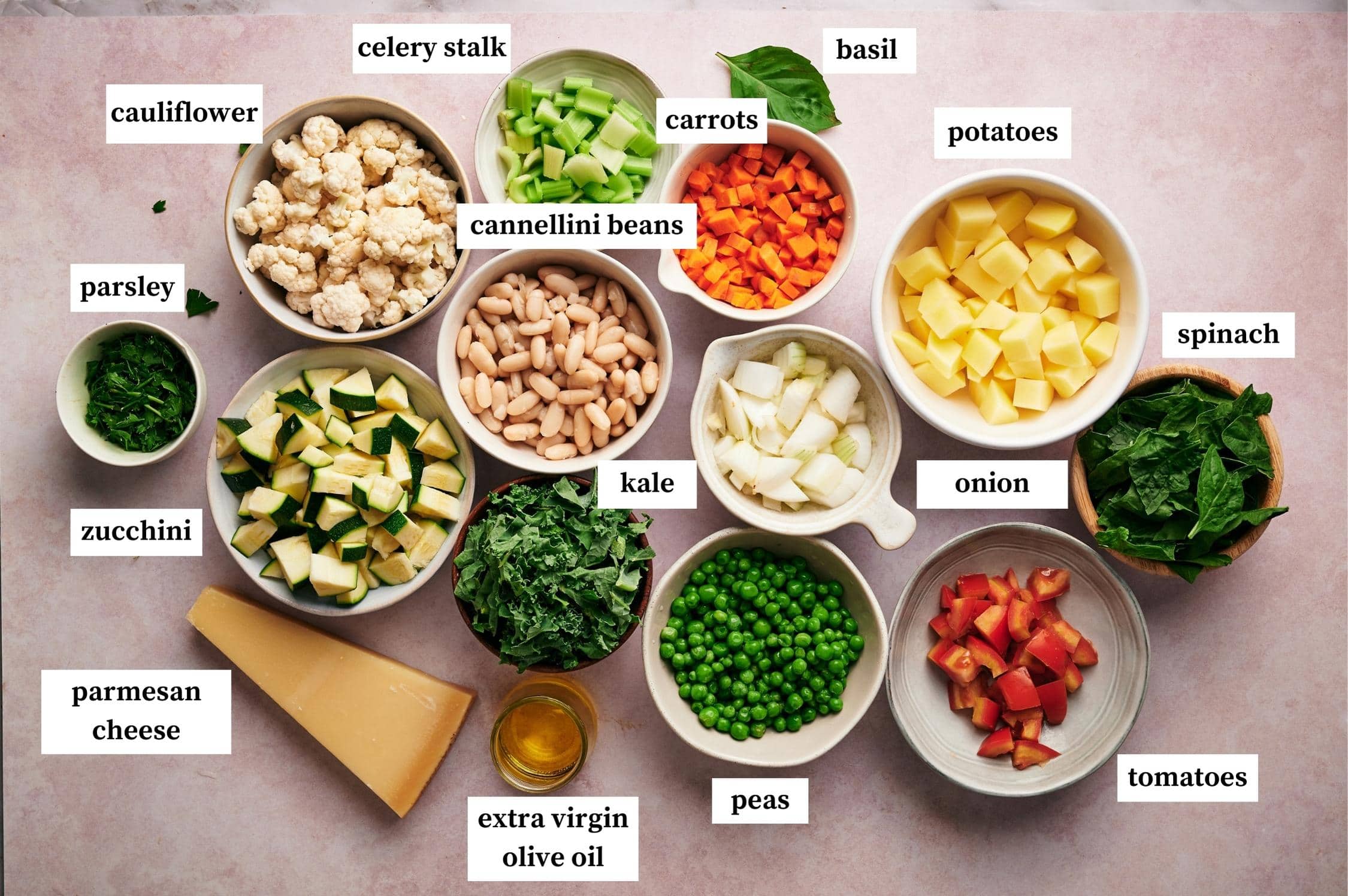 Ingredients for Minestrone soup: cauliflower, celeery stalk, carrots, basil, potatoes, spinach, onion, tomatoes, peas, kale, extra virgin olive oil, cannellini beans, parmesan cheese, zucchini and parsley.