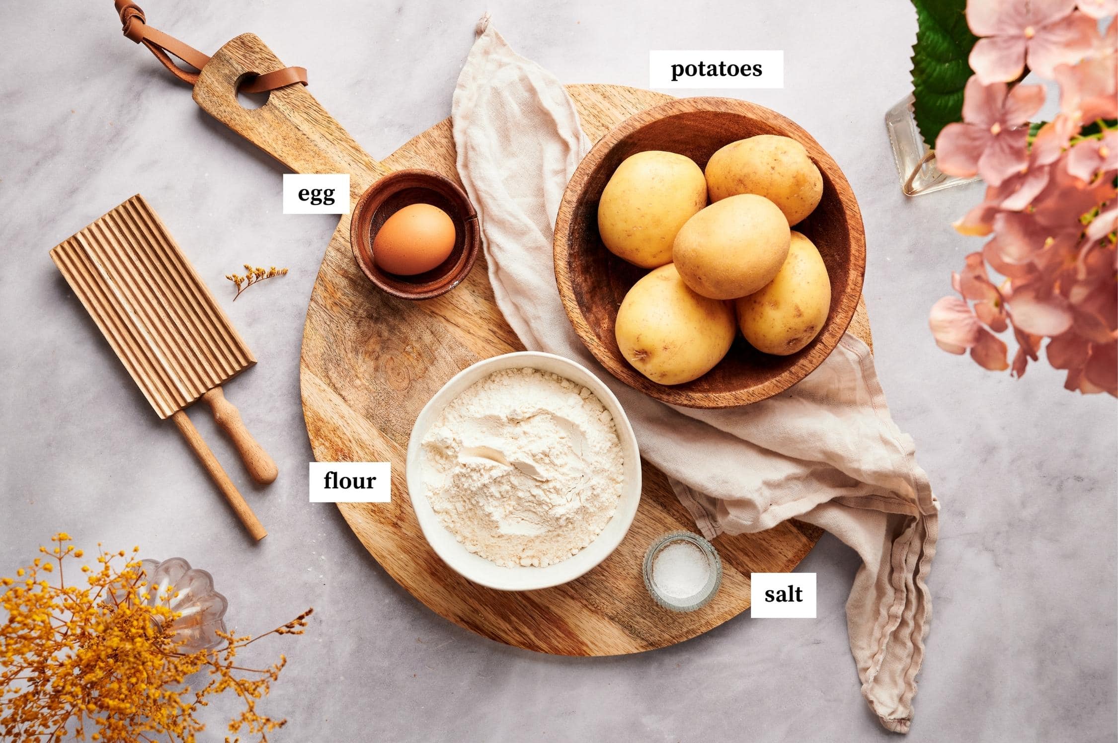 ingredients of potato gnocchi, potatoes in a wooden bowl, egg, bowl of flour, and salt, on top of a round chopping board