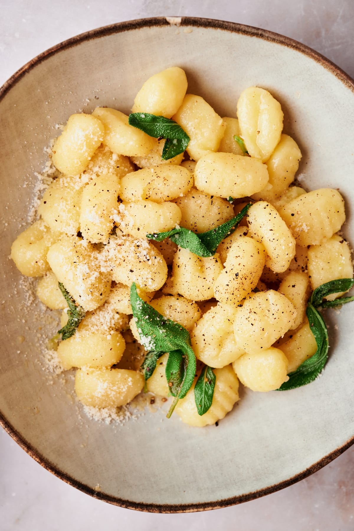 finished plate of potato gnocchi on a brown plate topped with sage, ground black pepper and parmesan cheese