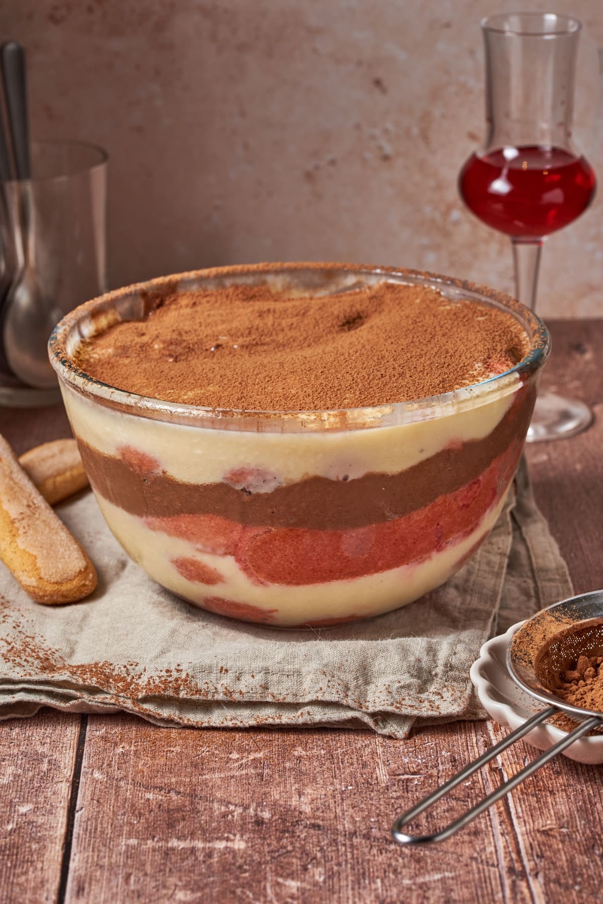 bowl of zuppa inglese (italian trifle) on a plate with a glass containing alchermes liqueur in the background